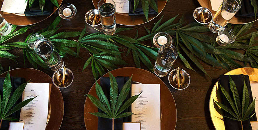 Overhead view of a cannabis-themed dining table with gold chargers, black napkins, menus, candles and glassware arranged on a dark wood table, decorated with large marijuana leaves as leafy centerpieces.