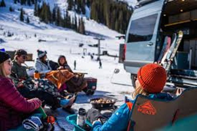 Cozy après-ski scene: a group in winter gear sitting by a camper van with skis propped up, enjoying hot drinks and a small fire on a sunny snowy mountain at a ski resort.