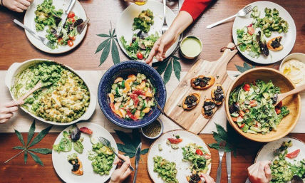Top-down view of a communal plant-based meal on a wooden table: salads, cheesy green pasta, mixed vegetable bowl, crostini, and decorative cannabis leaves.