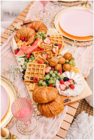 Outdoor summer brunch charcuterie board on a picnic table with flaky croissants, mini waffles topped with strawberries and raspberries, a brie wheel with berries, green grapes, watermelon slices, muffins and cheese cubes, styled with pink plates and a goblet.