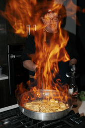 Cook flambés sliced bananas in a stainless-steel skillet over a gas stove, bright orange flames leaping up with a liquor bottle nearby in an indoor kitchen.