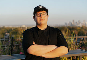 Confident chef in a black coat and Yankees cap with glasses, arms crossed on a rooftop patio overlooking a city skyline in golden-hour light.