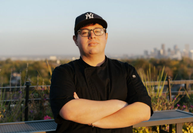 Rooftop portrait of a chef in a black coat and glasses with a dark baseball cap, arms crossed, city skyline and sunset in the background.