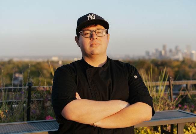 Confident chef in a black jacket and navy baseball cap with glasses stands on a rooftop terrace at golden hour, arms crossed with a blurred urban skyline in the background.