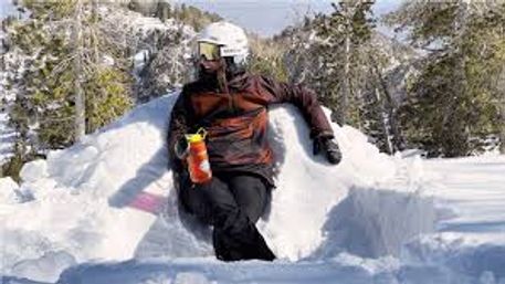 Person in ski helmet and goggles relaxing on a carved snow seat, holding an orange water bottle on a sunny alpine slope with snow-covered trees.
