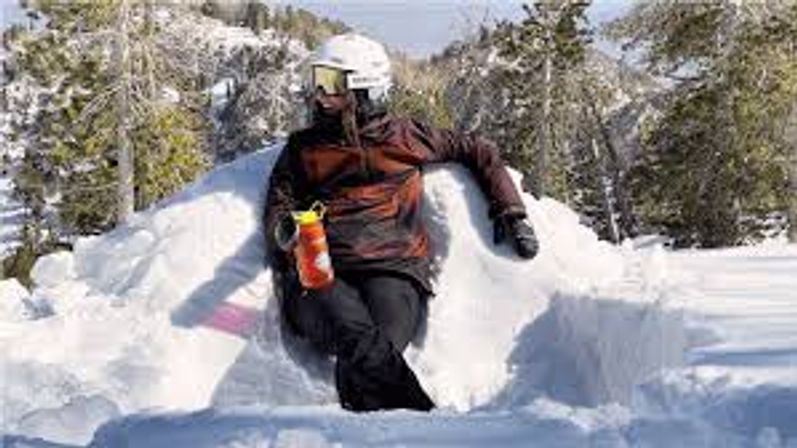 Person in ski helmet and goggles relaxing on a carved snow seat, holding an orange water bottle on a sunny alpine slope with snow-covered trees.