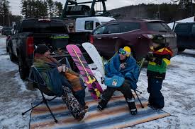 Group of snowboarders tailgating in a snowy ski-area parking lot, sitting in folding chairs beside trucks and colorful snowboards wearing winter gear and goggles
