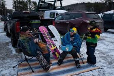 Group of snowboarders tailgating in a snowy ski-area parking lot, sitting in folding chairs beside trucks and colorful snowboards wearing winter gear and goggles