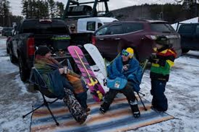 Group of snowboarders tailgating in a snowy ski-area parking lot, sitting in folding chairs beside trucks and colorful snowboards wearing winter gear and goggles