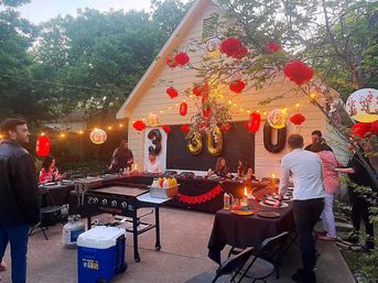 Backyard 30th birthday party at a garage patio with string lights, red paper lanterns and gold 30 balloons, tables with black linens, an outdoor griddle and guests mingling under trees.