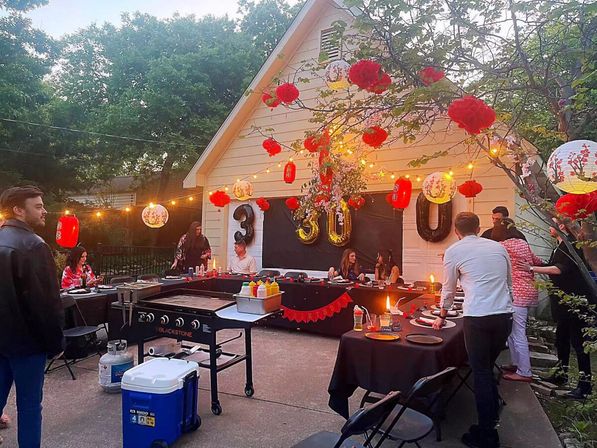 Backyard 30th birthday party at a garage patio with string lights, red paper lanterns and gold 30 balloons, tables with black linens, an outdoor griddle and guests mingling under trees.
