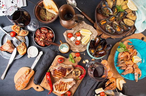Top-down view of a tapas-style seafood and grill spread on a dark table: mussels, prawns, grilled chops, bruschetta, roasted tomatoes, crusty bread and glasses of red wine.