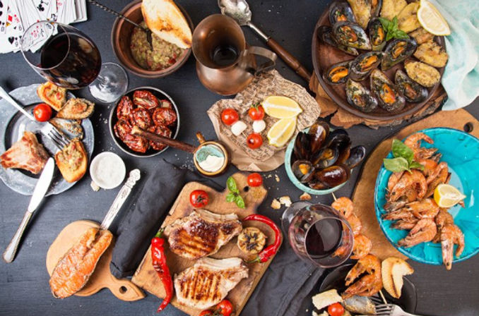 Top-down view of a tapas-style seafood and grill spread on a dark table: mussels, prawns, grilled chops, bruschetta, roasted tomatoes, crusty bread and glasses of red wine.