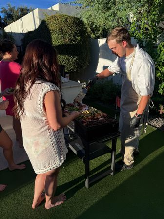 Backyard barbecue scene with a cook in an apron and gloves serving grilled meats and vegetables from a flat-top grill to a barefoot guest in a white cover-up on artificial turf during a sunny garden party.
