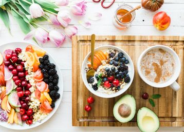 Sunlit overhead healthy breakfast: yogurt and granola bowl topped with blueberries, blackberries, cherries and sliced peach, avocado halves, frothy coffee, colorful fruit platter, honey jar and pink tulips on a wooden tray.