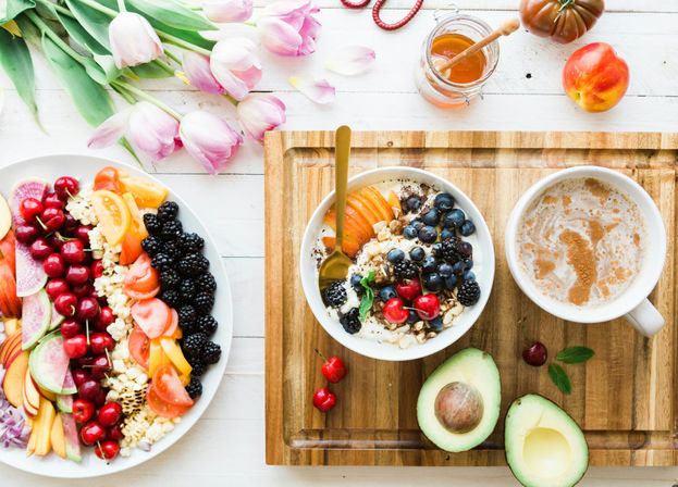 Sunlit overhead healthy breakfast: yogurt and granola bowl topped with blueberries, blackberries, cherries and sliced peach, avocado halves, frothy coffee, colorful fruit platter, honey jar and pink tulips on a wooden tray.