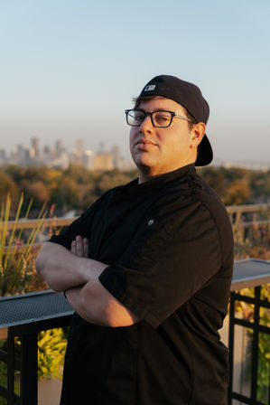 Rooftop portrait of a chef in a black jacket and backward baseball cap, arms crossed and wearing glasses at golden hour with a blurred city skyline and greenery in the background.