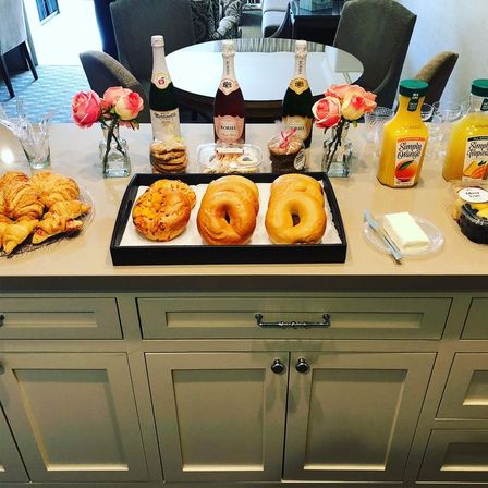 Bright kitchen island brunch spread with three bagels, pastries and croissants on trays, bottles of sparkling beverage, pitchers of orange juice, plates of butter and fruit, cookies and small vases of pink roses — casual breakfast buffet scene.