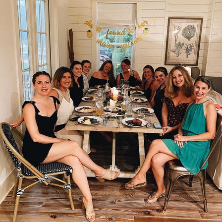 Group of women smiling at a celebratory dinner around a rustic wooden table in a shiplap dining room, lemon-themed banner overhead, plated meals, wine glasses and candles.