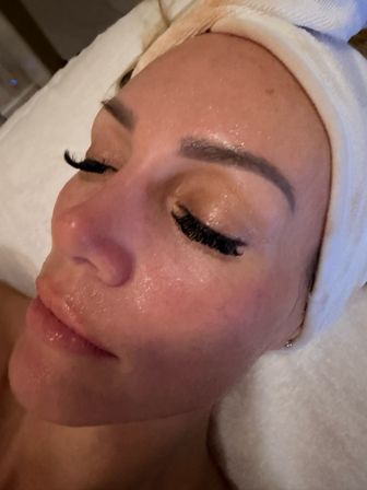 Close-up spa facial of a relaxed person wearing a towel headband, glossy moisturized skin, sculpted brows and dramatic eyelash extensions.