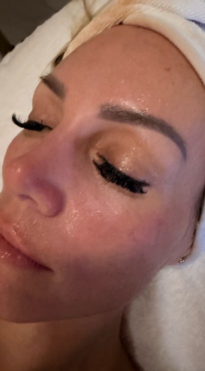Close-up spa facial of a relaxed person wearing a towel headband, glossy moisturized skin, sculpted brows and dramatic eyelash extensions.