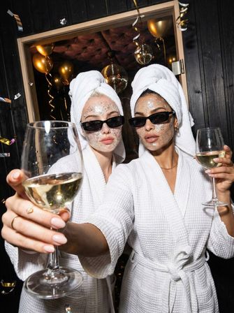 Two women in white spa robes and towel turbans wearing glitter face masks and black sunglasses, toasting white wine amid gold balloons, disco ball and confetti for a playful spa-party vibe.