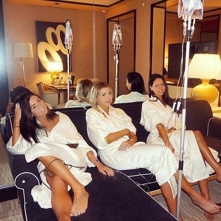Three women in white spa robes lounging on a hotel-suite sofa, each receiving IV therapy drips and smiling in a cozy, lamp-lit wellness setting