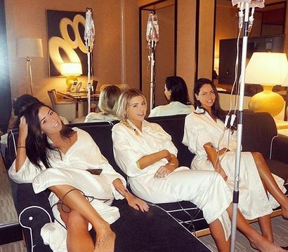 Three women in white spa robes lounging on a hotel-suite sofa, each receiving IV therapy drips and smiling in a cozy, lamp-lit wellness setting
