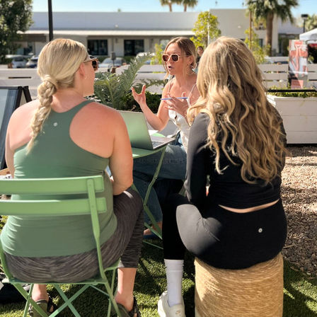 Three women in athletic wear chatting at a small outdoor patio table — one sunglasses-wearing woman gestures while speaking over a laptop, two others sit with backs to camera on green chairs and a woven stool in a sunny, palm-lined courtyard.