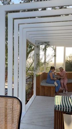 Modern white pergola on an elevated outdoor deck with wooden slatted benches and green-and-white striped cushions; two people chatting while enjoying a sunlit valley view at sunset.