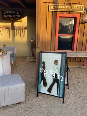 Rustic Arizona storefront with a wooden sheriff’s office sign, vintage blacksmith tools on the wall, and a playful A-frame sign promoting a cosmic coaching service in a sandy courtyard.