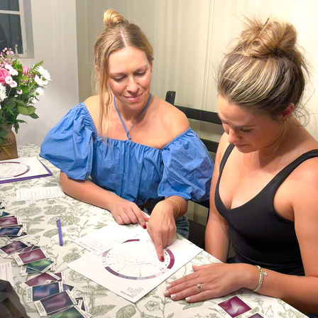 Two women at a home dining table examining an astrological birth chart as one points to the zodiac wheel; a spread of oracle/tarot cards and a vase of fresh flowers nearby.