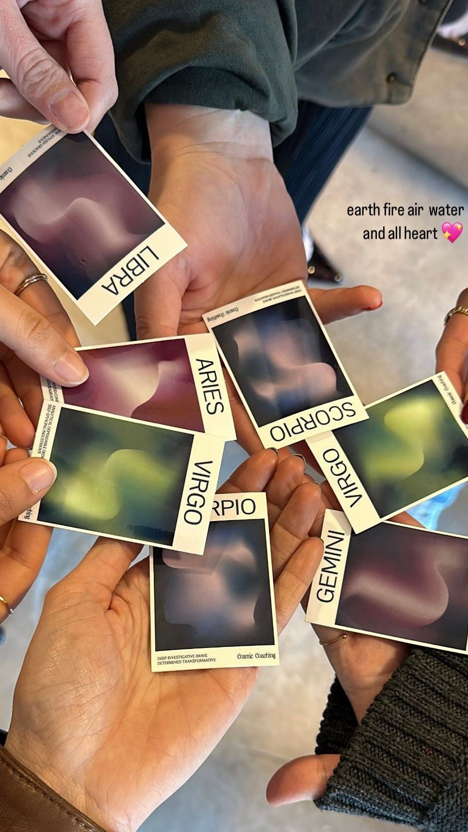 Close-up of hands holding colorful zodiac cards labeled Libra, Aries, Virgo, Scorpio and Gemini with abstract swirls and text overlay “earth fire air water and all heart,” cozy group astrology reading