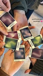 Close-up of hands holding colorful zodiac cards labeled Libra, Aries, Virgo, Scorpio and Gemini with abstract swirls and text overlay “earth fire air water and all heart,” cozy group astrology reading