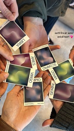 Close-up of hands holding colorful zodiac cards labeled Libra, Aries, Virgo, Scorpio and Gemini with abstract swirls and text overlay “earth fire air water and all heart,” cozy group astrology reading