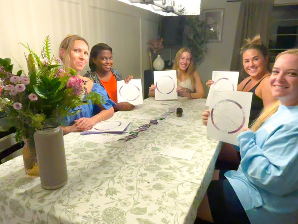 Five friends gathered around a home dining table, smiling and holding circular astrology/star charts with a floral centerpiece in the foreground.