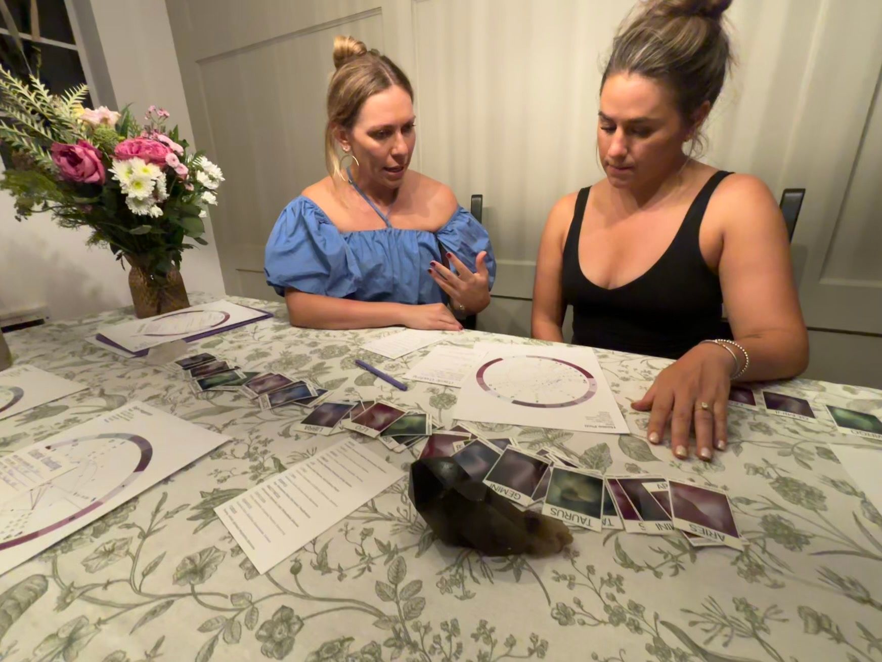 Two women at a cozy home dining table doing a tarot-style card spread with an astrology chart and dark crystal on a floral tablecloth, vase of flowers nearby.
