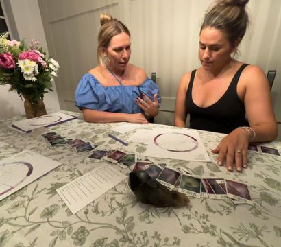Two women at a cozy home dining table doing a tarot-style card spread with an astrology chart and dark crystal on a floral tablecloth, vase of flowers nearby.