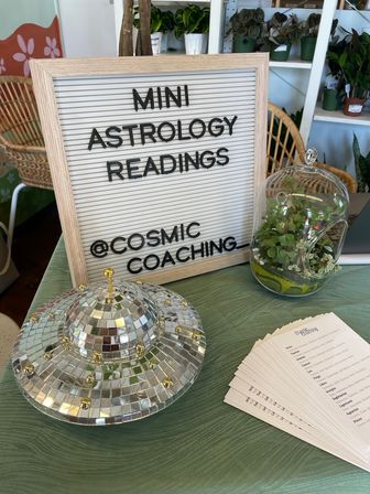 Letter board reading "MINI ASTROLOGY READINGS" displayed on a table with a mirrored disco‑UFO decoration, glass succulent terrarium, and stacked astrology cards in a bright indoor plant shop setting.