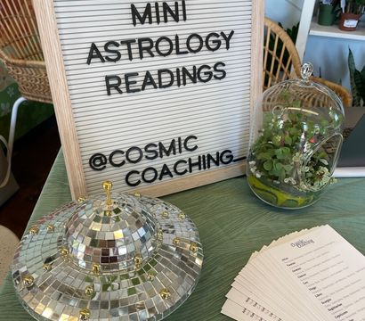 Letter board reading "MINI ASTROLOGY READINGS" displayed on a table with a mirrored disco‑UFO decoration, glass succulent terrarium, and stacked astrology cards in a bright indoor plant shop setting.