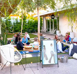 Sunlit courtyard wellness workshop with people seated on yoga mats on artificial grass, chatting in small groups under trees beside patio chairs and an A-frame sign — outdoor group coaching and community yoga session.