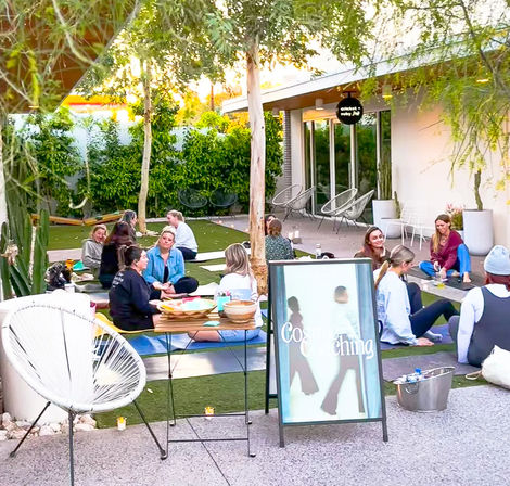 Sunlit courtyard wellness workshop with people seated on yoga mats on artificial grass, chatting in small groups under trees beside patio chairs and an A-frame sign — outdoor group coaching and community yoga session.