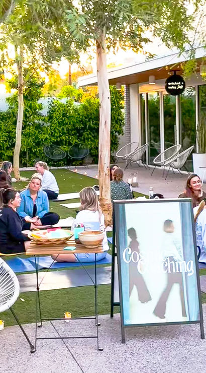 Sunlit courtyard wellness workshop with people seated on yoga mats on artificial grass, chatting in small groups under trees beside patio chairs and an A-frame sign — outdoor group coaching and community yoga session.