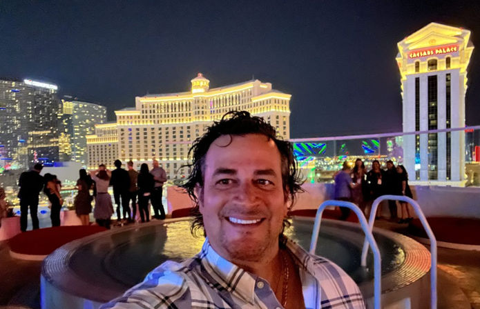 Person smiling for a selfie beside a rooftop hot tub at night with an illuminated Las Vegas Strip skyline, glowing hotel towers and groups of people mingling on the terrace.