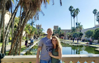Smiling couple posing at a stone railing overlooking a long reflecting pool lined with palm trees and Spanish‑style buildings on a sunny day.