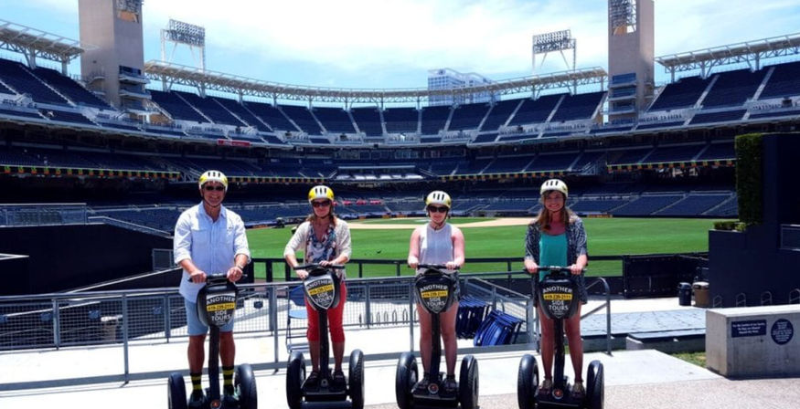 Four helmeted riders on Segways lined up with a green baseball field and empty stadium seats behind them during a sunny stadium tour.