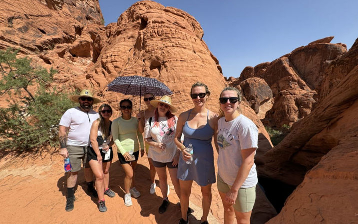 Group of seven hikers posing on a sunlit red sandstone canyon trail with towering red rock formations and blue sky; wearing sun hats, sunglasses and light hiking clothes, one holds a patterned umbrella and several hold water bottles.