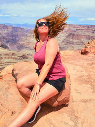Woman in sunglasses sitting on red rock at an Arizona canyon overlook, hair blowing in the wind with layered canyon and river below on a sunny day.