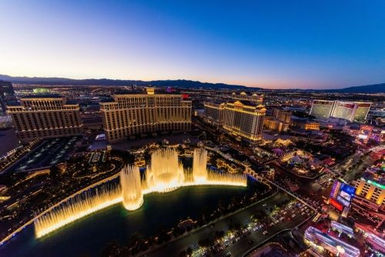 Aerial dusk view of the Las Vegas Strip with a dazzling illuminated fountain show in a curved lake, glowing resort hotels and neon-lit streets stretching into the desert.