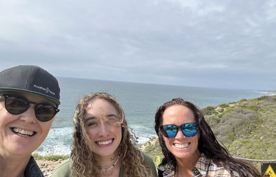 Three smiling people take a selfie on a rocky coastal bluff overlooking the ocean and waves, wind-tousled hair, sunglasses, and an overcast sky.